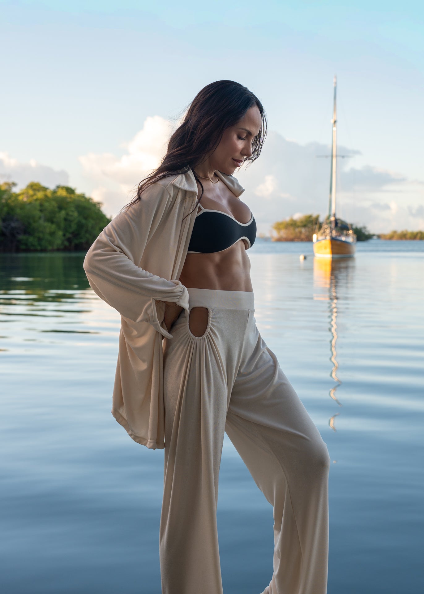 Woman in a black bikini top and beige pants standing by a body of water with a sailboat in the background.