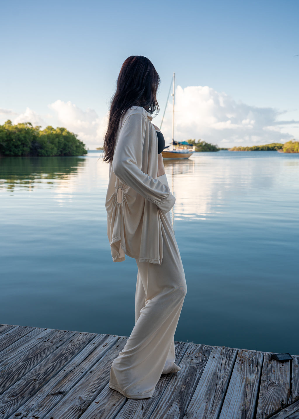 Woman in a beige outfit standing on a wooden dock by a calm body of water with a sailboat in the background.