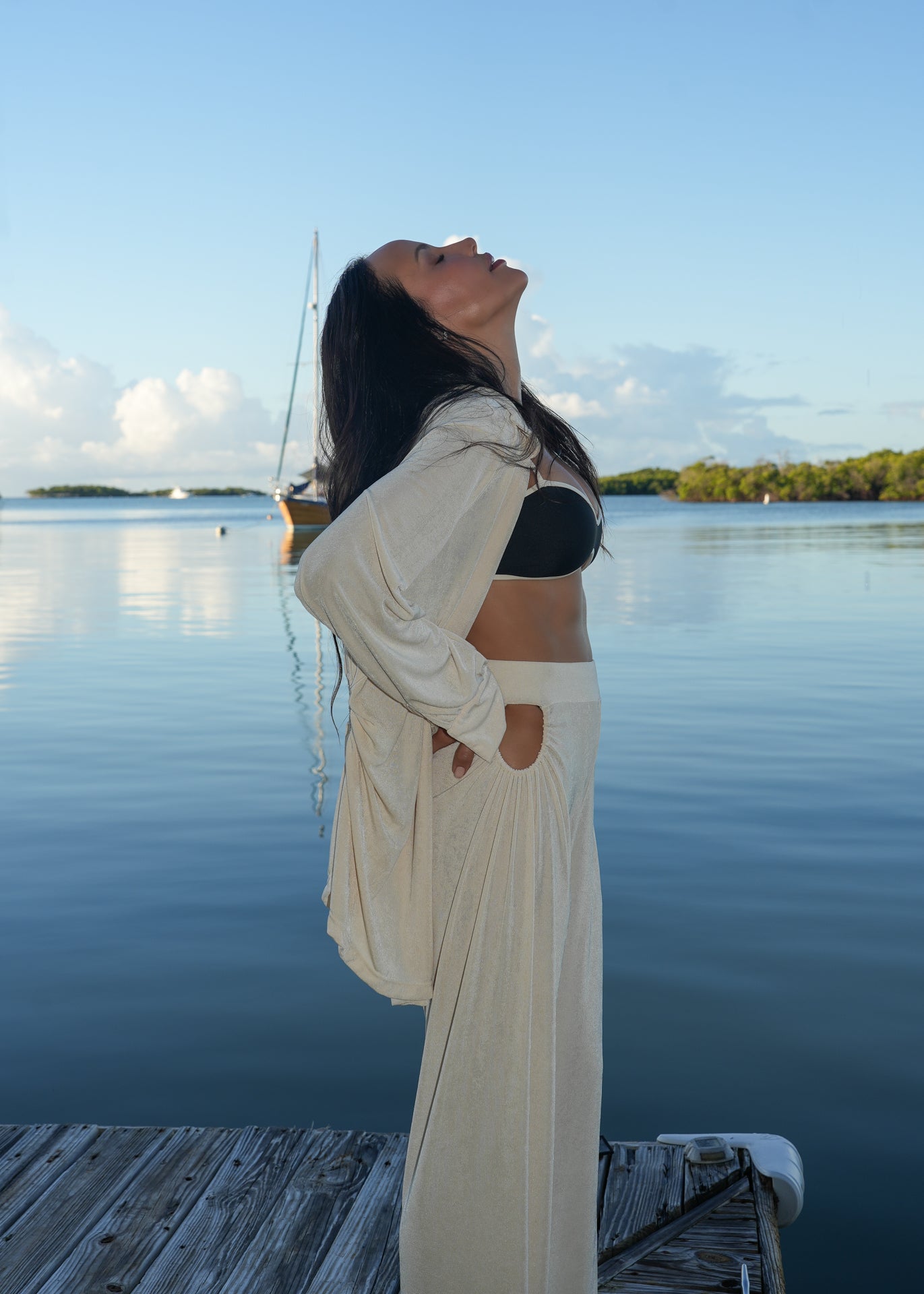 Woman in a white outfit standing on a dock by a body of water with a sailboat in the background.